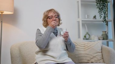 Kanepede oturan yaşlı bir kadın, suyla birlikte bir hap alıyor. Evde bir hastalık, ağrı ya da kronik durumu tedavi ediyor gibi görünüyor..