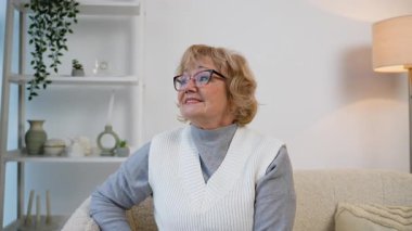 Gözlüklü, rahat bir koltukta oturan, gülümseyen ve kameraya bakan yaşlı bir kadının yakın çekimi. Mutlu, pozitif ve evinde mutlu görünüyor..