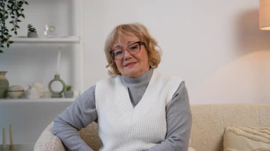 Gözlüklü yaşlı bir kadın kanepeye oturmuş üzgün ve düşünceli görünüyor. Derin düşüncelere dalmış gibi görünüyor, duygusal bir şeyi yansıtıyor..