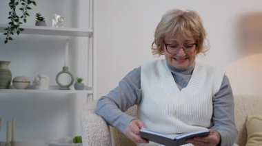 Gözlüklü gülümseyen yaşlı kadın, güzel dekore edilmiş evinde rahat bir kanepede otururken kurgu kitapları okumaktan zevk alır..