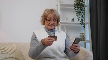 Yaşlı bir kadın rahat bir koltukta oturuyor, elinde akıllı telefon ve kredi kartı var. Çevrimiçi alışveriş yapıyor ya da mali işler yönetiyor gibi görünüyor..