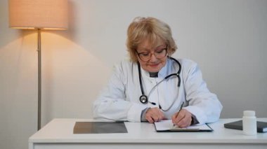 Steteskoplu beyaz önlüklü yaşlı kadın doktor masasına notlar yazıyor. Profesyonel, arkadaş canlısı ve kendine güvenen birine benziyor..