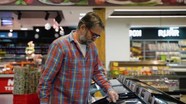 Ekose gömlekli ve eldivenli yaşlı bir adam süpermarkette donmuş kırmızı balığı inceliyor. Dikkatlice deniz mahsulleri seçerken bir dondurucu sergisi açıyor..