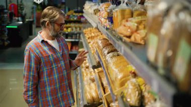 Ekose gömlekli yaşlı bir adam bir süpermarketin fırın bölümünden dikkatlice ekmek seçer. Raftaki farklı paketlenmiş somunları inceliyor..