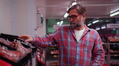 Ekose gömlekli yaşlı bir adam soğutmalı marketlerde paketlenmiş et seçiyor. Satın almadan önce indirimli ürünleri dikkatle kontrol eder..