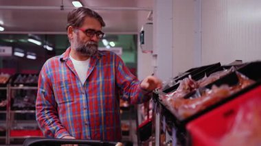 Ekose gömlekli yaşlı bir adam bir süpermarketin et bölümüne bakıyor. Dondurucu koridorda bir alışveriş arabasını iterken paketlenmiş eti dikkatle seçiyor..