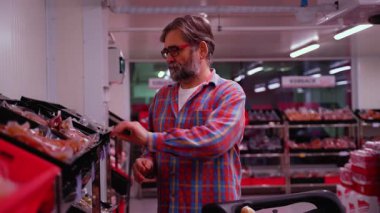Ekose gömlekli yaşlı bir adam bir süpermarketin et bölümüne bakıyor, bir alışveriş arabasını iterken paketlenmiş bifteği dikkatlice seçiyor..