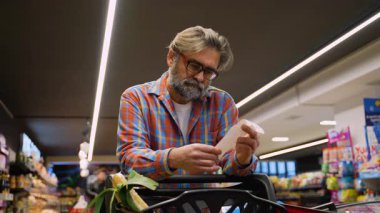 Ekose gömlekli yaşlı bir adam markette alışveriş arabasının yanında durup alışveriş ve harcamalarını kontrol ederken alışveriş makbuzunu yakından inceliyor..