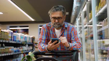Ekose gömlekli yaşlı bir adam marketin koridorunda duruyor, alışveriş arabasına yaslanıyor ve akıllı telefonundan aldığı yiyecekleri kontrol ediyor..
