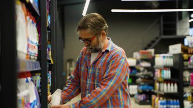 Ekose gömlekli yaşlı bir adam süpermarket temizlik koridorunda bir şişe deterjanı inceliyor. İhtiyaçları için doğru temizlik ürününü dikkatle seçer..