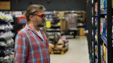 Ekose gömlekli yaşlı bir adam süpermarketteki temizlik ürünleri reyonuna bakıyor ve ihtiyaçları için en iyi deterjanı dikkatlice seçiyor..