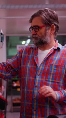 Ekose gömlekli yaşlı bir adam soğutmalı marketlerde paketlenmiş et seçiyor. Satın almadan önce indirimli ürünleri dikkatle kontrol eder..