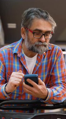 Ekose gömlekli yaşlı bir adam marketin koridorunda duruyor, alışveriş arabasına yaslanıyor ve akıllı telefonundan aldığı yiyecekleri kontrol ediyor..
