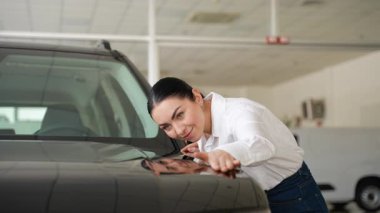 Beyaz gömlekli ve kotlu gülümseyen kadın bir galeride bir arabanın yüzeyini yakından inceliyor..