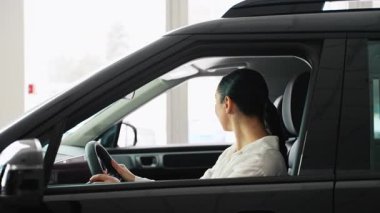 Beyaz gömlekli bir kadın, satış mağazasının içindeki siyah bir arabanın sürücü koltuğunda oturuyor. Satın alma kararı vermeden önce iç mekanı dikkatlice inceliyor..