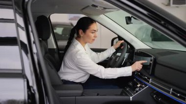 Beyaz gömlekli bir kadın, satış mağazasının içindeki siyah bir arabanın sürücü koltuğunda oturuyor. Satın almadan önce dokunmatik ekran ve gösterge paneliyle etkileşime giriyor..