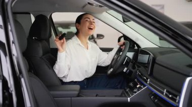 Beyaz gömlekli neşeli bir kadın yeni siyah arabasının şoför koltuğunda oturuyor. Anahtarları elinde tutuyor ve bir galeride alışverişini kutluyor..