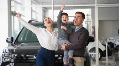 Küçük bir çocukla neşeli bir aile, bir galerinin içinde, yeni siyah arabalarının önünde duruyor. Başarılı alışverişlerini alkışlıyor ve kutluyorlar..