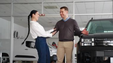 Neşeli bir çift, bir galeri galerisinde yeni siyah arabalarının yanında duruyor. Gülümsüyor ve başarılı alışverişlerini ve yeni ve heyecan verici yolculuklarını kutluyorlar..