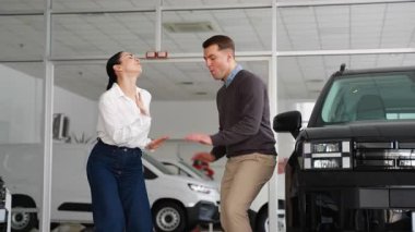 Neşeli bir çift, yeni siyah arabalarının yanında bir galeri galerisinde dans eder ve alışverişlerinin heyecanını ve mutluluğunu ifade eder..