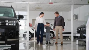Küçük bir çocukla gülümseyen bir aile bir araba galerisinde el ele yürüyor, araç seçeneklerini araştırıyor ve yeni bir araba seçme sürecinin tadını çıkarıyor..