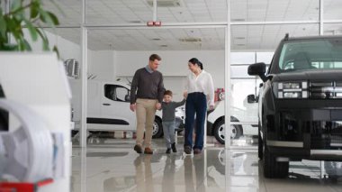 Küçük çocuklu neşeli bir aile bir araba galerisinde el ele yürür, farklı araç seçeneklerini araştırır ve yeni arabalarını birlikte seçerler..