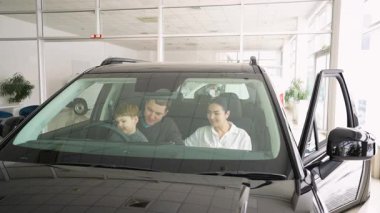 Küçük bir oğlanla mutlu bir aile bir galeride sağdan çekişli bir arabada oturur. Satın alma kararı vermeden önce içeriyi ve özellikleri kontrol eder..