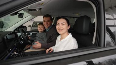 Gülümseyen bir aile, yeni satın aldıkları sağ şeritli arabalarının içinde yeni arabaları için heyecanlı bir şekilde oturuyor. Neşeli ve memnun müşteriler..