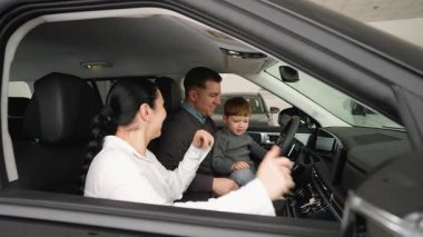 Neşeli bir aile, yeni satın aldıkları arabanın içinde bir galeride oturup satın almalarını kutluyor. Ebeveynler ve çocuklar modern araçtan birlikte hoşlanıyorlar..