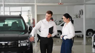 Beyaz gömlekli profesyonel bir araba satıcısı, parlak, modern bir galeride çalışan bir müşteriye siyah bir SUV 'nin özelliklerini açıklar..