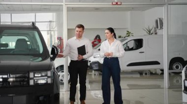 Beyaz gömlekli bir araba satıcısı modern bir galeride çalışan bir bayana özel indirimli siyah bir SUV sunar..