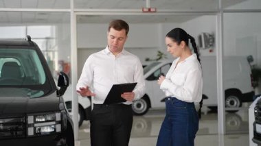 Bir araba satıcısı yeni bir aracın anahtarlarını galerinin içindeki bir kadın müşteriye verir. Satın alma ve araba özelliklerini tartışarak sonuçlandırır..