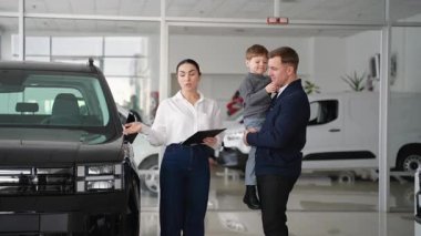 Bir kadın araba satıcısı, oğlunu bir galeride tutan bir babayla araç seçeneklerini tartışıyor ve doğru arabayı seçmelerine yardım ediyor..