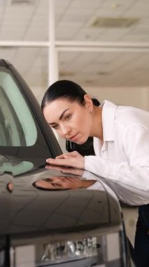 Beyaz gömlekli ve kotlu gülümseyen kadın bir galeride bir arabanın yüzeyini yakından inceliyor..