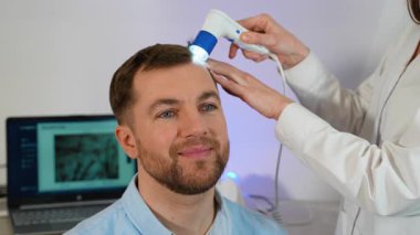 Trikoloğun biri klinikte dermatoskop kullanarak gülümseyen bir adamın kafa derisini inceliyor. Bir dizüstü bilgisayar büyütülmüş saç analizi sonuçlarını gösterir.