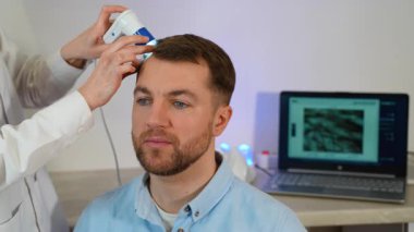 Erkek bir hasta, dermatoskop kullanan bir uzman tarafından, büyütülmüş saç analizi sonuçlarını gösteren bir dizüstü bilgisayarla kafa derisi muayenesinden geçirilir..