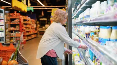 Gözlük takan yaşlı bir kadın süpermarketteki soğutulmuş bir raftan süt ürünleri seçiyor ve içi yiyecek dolu bir arabayı itiyor..