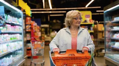 Gözlüklü ve alışveriş arabalı yaşlı bir kadın bir süpermarket koridorunda geziniyor, parlak mağaza ışıklarının altındaki raflardan taze sebzeler seçiyor..