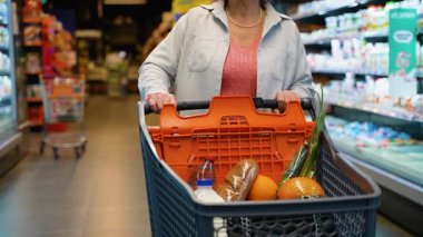 Son sınıf öğrencisi bir kadın marketin koridorunda süt, ekmek, portakal ve yeşil soğan gibi taze yiyeceklerle dolu bir alışveriş arabasını itiyor..