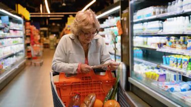 Gözlüklü yaşlı bir kadın makbuzunu incelerken içi taze sebze dolu alışveriş arabasını iyi aydınlatılmış bir süpermarket koridorunda itiyor..