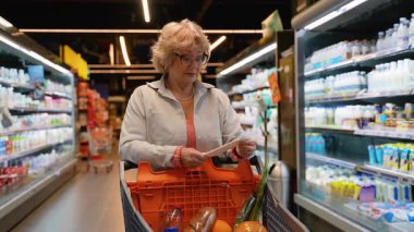 Gözlüklü yaşlı bir kadın alışveriş arabasının fişini incelerken süpermarket koridorunda içi taze yiyeceklerle dolu bir alışveriş arabasını itip harcamalarını kontrol ediyor..