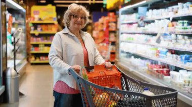 Gözlüklü ve alışveriş arabalı yaşlı bir kadın süpermarket koridorunda dolaşırken gülümsüyor, buzdolabından taze sebze seçiyor..
