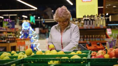 Gözlüklü ve açık renk ceketli yaşlı bir kadın marketten taze elma topluyor ve satın almak için en iyi meyveyi dikkatlice seçiyor..