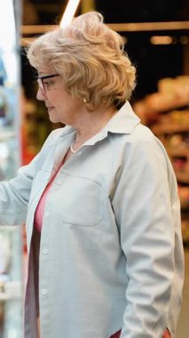 Gözlüklü yaşlı bir kadın marketteki soğutuculu bir raftan yoğurt seçerken içi yiyecek dolu bir alışveriş arabasını itiyor..