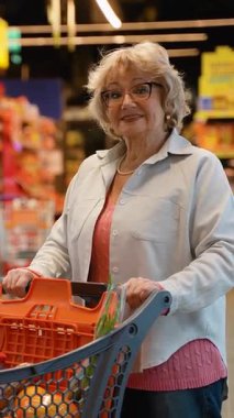 Gözlüklü ve alışveriş arabalı yaşlı bir kadın süpermarket koridorunda dolaşırken gülümsüyor, buzdolabından taze sebze seçiyor..