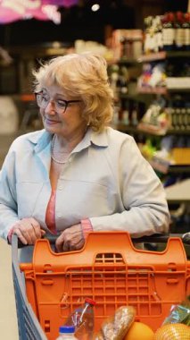 Gözlüklü yaşlı bir kadın süpermarketin soğutucu bölümüne bakarken bir alışveriş arabasını itiyor ve raflardan karbonatlı içecekler seçiyor..