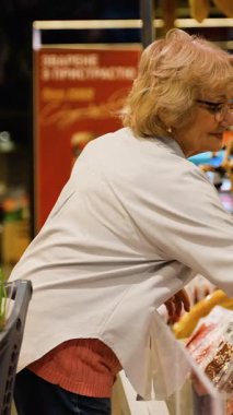 Gözlüklü ve alışveriş arabalı yaşlı bir kadın süpermarkette kaliteli et ürünleri seçen gurme sosisleri dikkatle inceliyor..