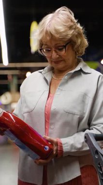 Yaşlı bir kadın alışveriş arabası bir mağaza rafından temizlik malzemesi seçer, ev kullanımı için doğru deterjanı dikkatle seçer..