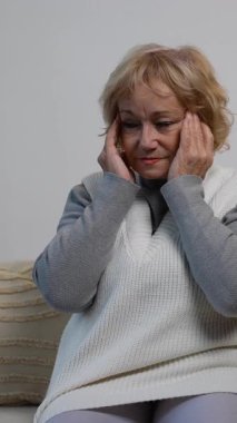 Rahat bir kanepede oturan, başı dertte olan yaşlı bir kadın, muhtemelen şiddetli bir baş ağrısı, migren ya da strese bağlı bir rahatsızlık yaşıyor. İlaç ve su yakında..