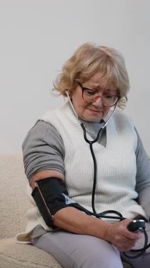 Kanepede oturan yaşlı bir kadın manuel sfenometre ve stetoskop kullanarak kan basıncını ölçüyor. Evde sağlığını gözetiyor..